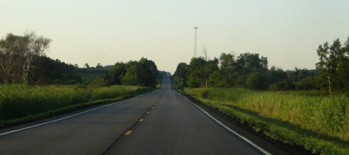 A straight, empty road stretches into the distance, flanked by green fields and trees under a clear sky.