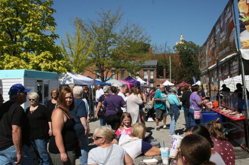 A bustling outdoor market scene with people browsing stalls, enjoying food, and socializing under a clear blue sky.