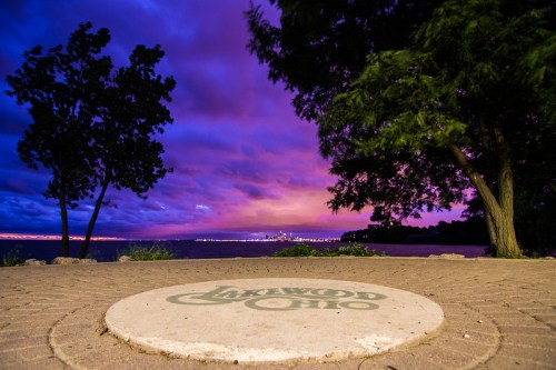 A scenic view of a lake at sunset, with vibrant purple clouds and a circular stone featuring "Lakewood, Ohio."