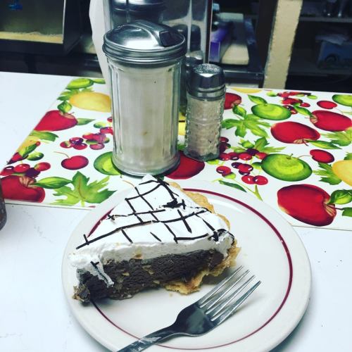 A slice of chocolate cream pie with whipped cream on a plate, next to a sugar dispenser and salt shaker on a colorful tablecloth.