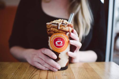 A person holding a tall glass of chocolate milkshake topped with nuts and drizzled chocolate sauce.