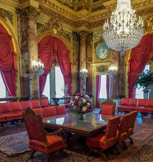 Luxurious room with ornate decor, red drapes, chandeliers, and a large table surrounded by elegant chairs.
