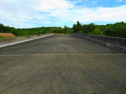 A wide, empty concrete surface with a slight incline, surrounded by greenery and under a cloudy sky.