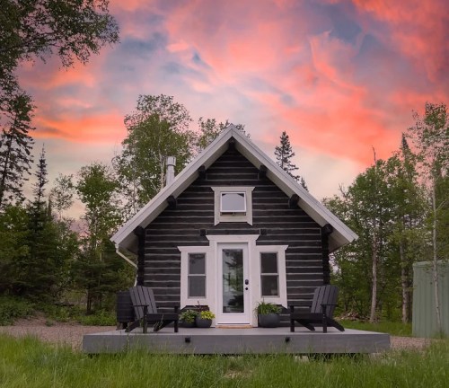 A cozy black log cabin with a white door, surrounded by greenery and a colorful sunset sky.