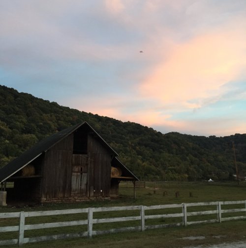 A rustic barn sits in a green field, surrounded by hills under a colorful sunset sky.
