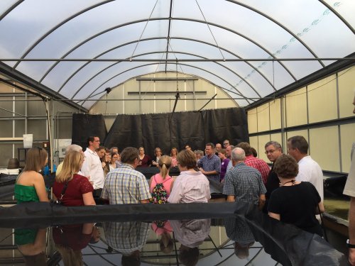 A group of people gathered inside a greenhouse, listening to a speaker near large tanks.