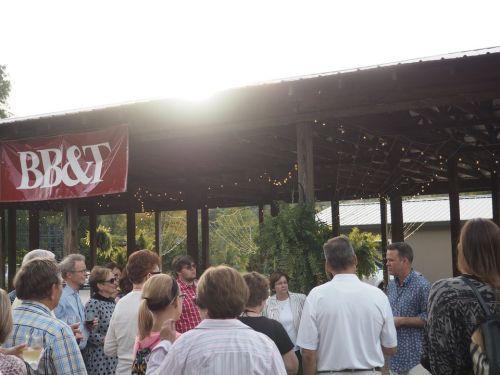 A group of people gathered under a covered outdoor space, listening to a speaker near a BB&T banner at sunset.