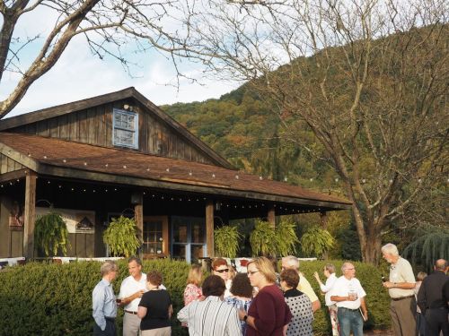 A gathering of people outside a rustic building with mountain views, surrounded by trees and festive decorations.