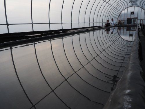 A greenhouse interior with a long, reflective water surface and two people seated at the far end.