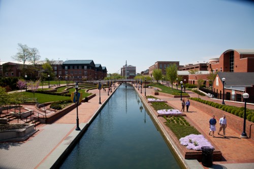A scenic view of a canal lined with trees and flowers, with people walking along pathways in a vibrant urban area.