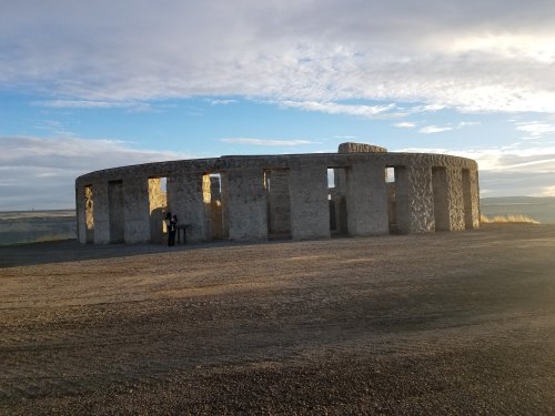 A stone structure resembling Stonehenge, set against a cloudy sky with two people standing nearby.