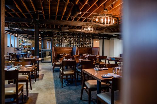 A modern restaurant interior featuring wooden tables, chairs, and a wine rack in a cozy, well-lit atmosphere.