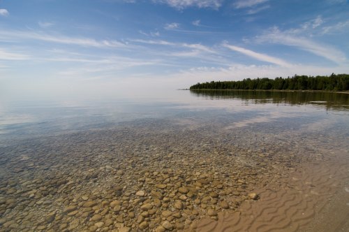 Calm, clear water reveals a rocky bottom, with a forested shoreline under a blue sky with wispy clouds.