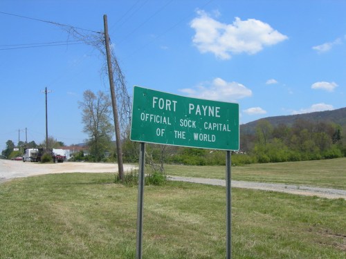 Green sign reading "Fort Payne, Official Sock Capital of the World" with a clear blue sky and grassy field in the background.