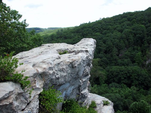 A rocky cliff edge overlooking a lush green valley and distant hills under a cloudy sky.