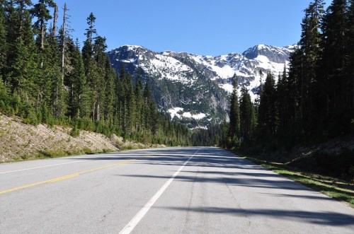 A clear road stretches through a forested area, with snow-capped mountains in the background under a blue sky.