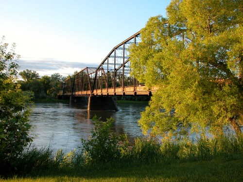 A historic bridge spans a river, surrounded by lush greenery and a clear blue sky.