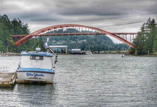 A boat named "Blue Yonder" in the foreground with a large orange bridge and trees in the background under a cloudy sky.