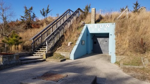 A blue entrance to a bunker with stairs leading up, surrounded by grass and trees under a clear blue sky.