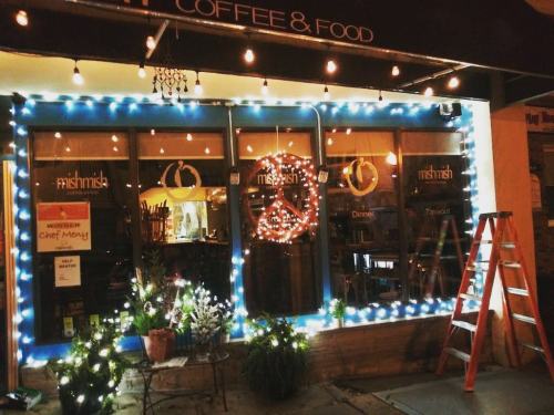 A cozy café with blue trim, festive lights, and a peace sign decoration in the window, featuring a ladder outside.