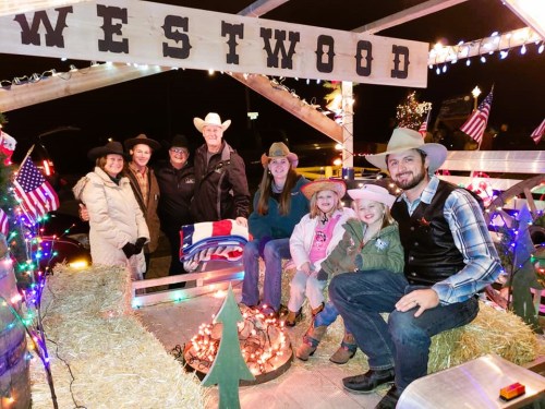A group of people in cowboy hats poses on a decorated float with lights and hay bales, celebrating a festive event.
