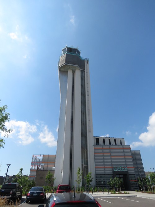 A tall, modern building with a glass observation deck against a blue sky, surrounded by parked cars and greenery.