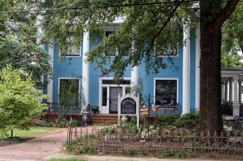 A blue two-story house with white columns, surrounded by greenery and a decorative fence.