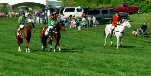 Three riders on horseback in colorful attire, with spectators and vehicles in the background on a grassy field.