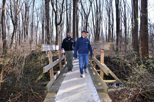 Two men walk across a wooden bridge in a wooded area, surrounded by bare trees and underbrush.
