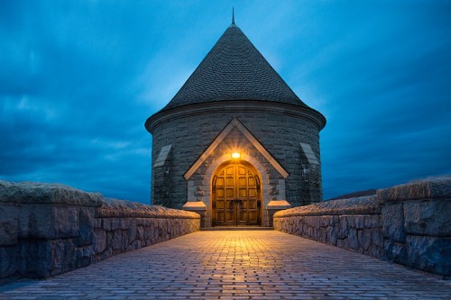 A stone tower with a conical roof, illuminated by a warm light, set against a twilight sky.