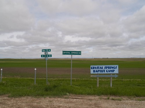 Road signs for Crystal Springs and Crystal Springs Baptist Camp in a grassy, open landscape under a cloudy sky.