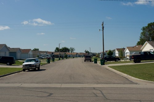 A quiet suburban street lined with houses and green trash bins on either side under a clear blue sky.
