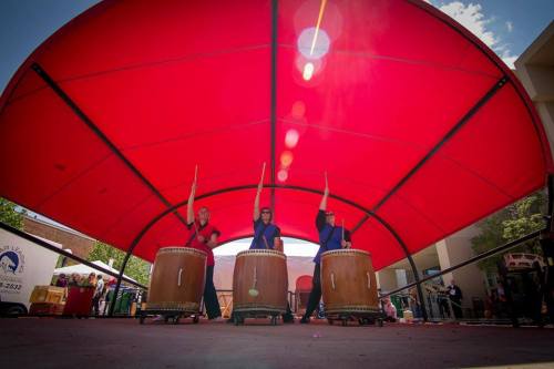 Three drummers perform under a large red canopy, raising their sticks in unison, with vibrant energy and focus.