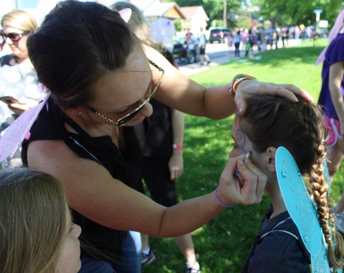 A woman paints a design on a girl's face at an outdoor event, with children and adults in the background.