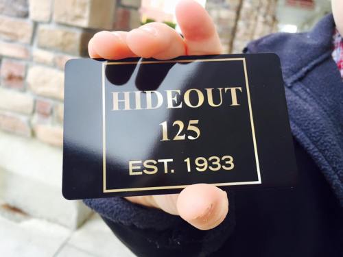 A person holds a black card that reads "HIDEOUT 125 EST. 1933" against a stone wall background.