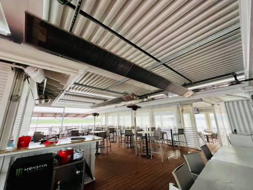 A spacious outdoor dining area with metal tables, chairs, and overhead fans, featuring a wooden floor and large windows.