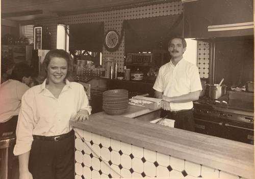 A black-and-white photo of a smiling woman and man behind a restaurant counter, with kitchen equipment in the background.