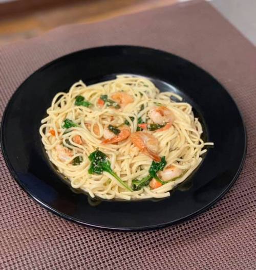 A black plate filled with shrimp pasta, garnished with spinach and herbs, on a textured brown surface.
