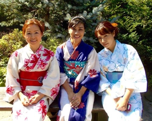 Three women in colorful kimonos sit together outdoors, surrounded by greenery and trees.