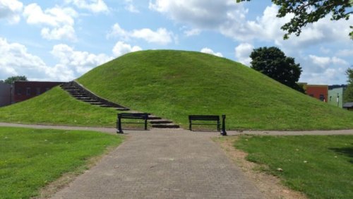 A grassy mound with stone steps leading up, surrounded by a park and blue sky with clouds. Benches are visible in front.
