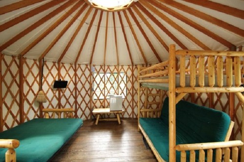 Interior of a yurt featuring wooden furniture, a bunk bed, and a cozy seating area with green cushions.