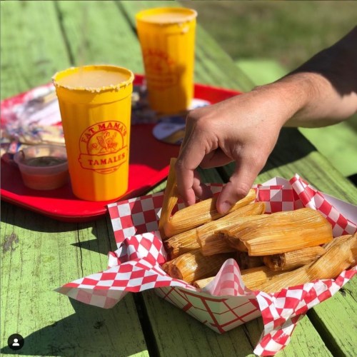 A hand reaching for tamales in a basket, with yellow cups and a red tray in the background on a green table.