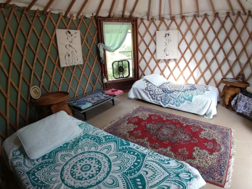 Cozy interior of a yurt with two beds, decorative rugs, and artwork on the walls. Natural light from a window.