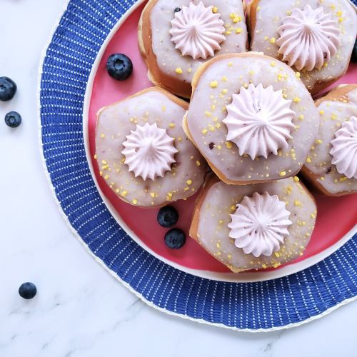 A plate of octagonal donuts with pink frosting, decorative swirls, and yellow sprinkles, surrounded by blueberries.