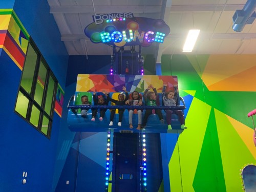 A group of children excitedly waving while seated on a colorful ride at an indoor play center.