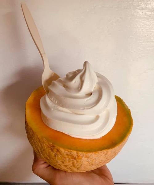 A hand holding a halved melon filled with soft-serve ice cream and a spoon on top.