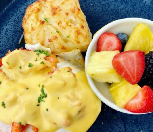 A plate featuring a seafood dish with hollandaise sauce, hash brown, and a bowl of mixed fruit including strawberries and pineapple.