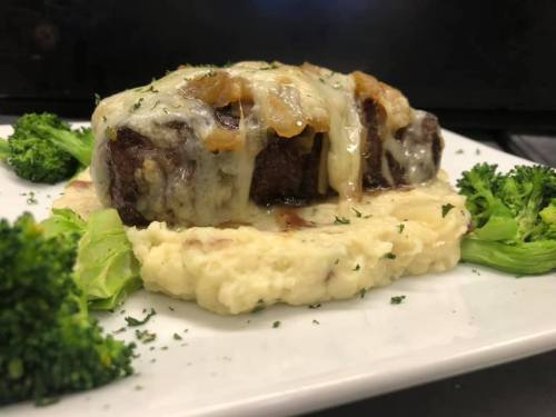 A plated dish featuring tender meat topped with melted cheese and onions, served with creamy mashed potatoes and broccoli.