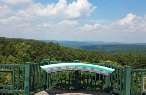 A scenic overlook with a green railing, featuring informational panels and a panoramic view of rolling hills and blue skies.