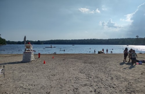 A sandy beach with a lifeguard tower, people swimming in the lake, and a cloudy sky in the background.